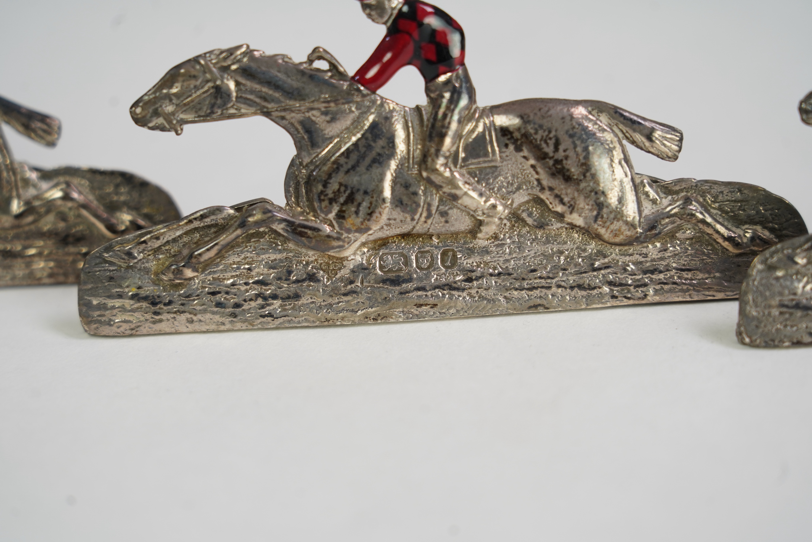 A set of four Elizabeth II silver and enamel menu holders, by Garrard & Co, each modelled as a racehorse and jockey with enamelled vest
